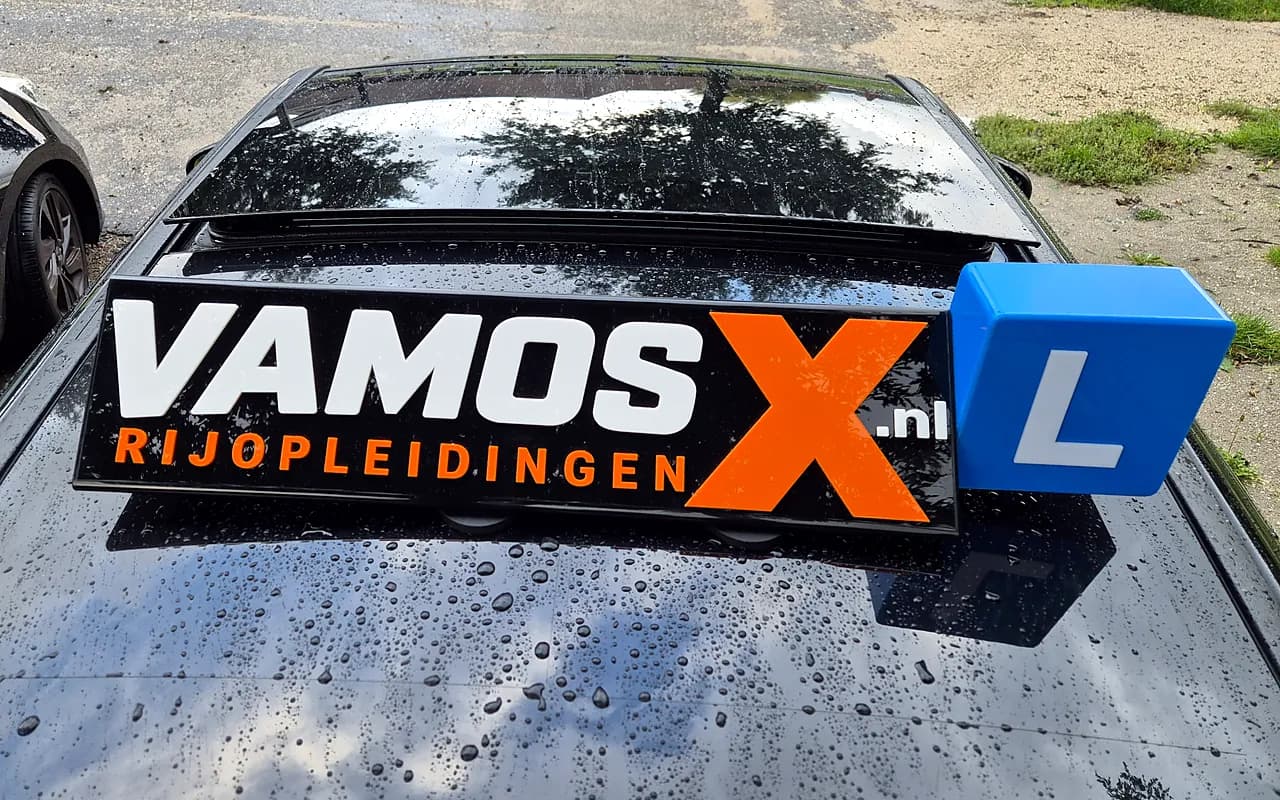 Het VamosX Rijopleidingen-dakbord met L-blok op het dak van de zwarte Mercedes