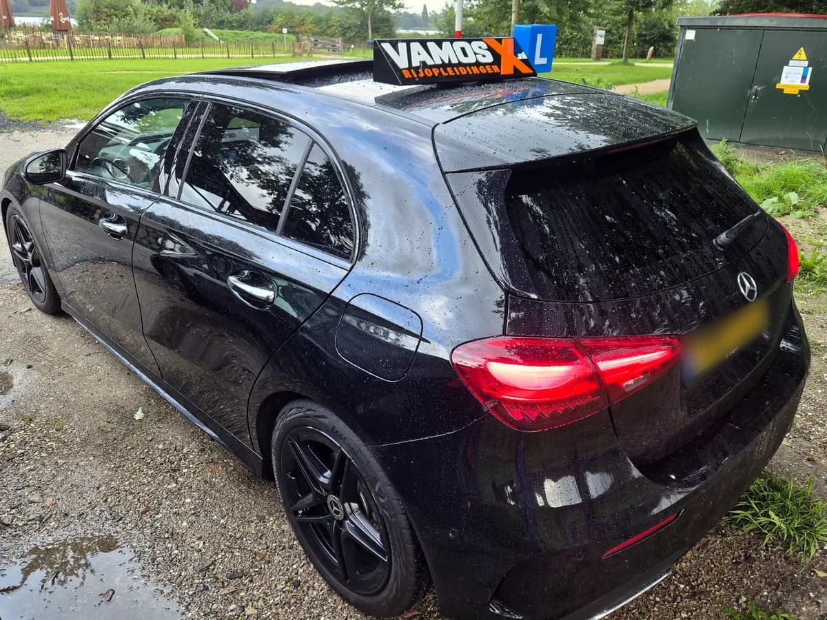 Onze zwarte Mercedes-Benz A180 automaat-lesauto van schuin achter, met het VamosX Rijopleidingen-dakbord en L-blok op het dak