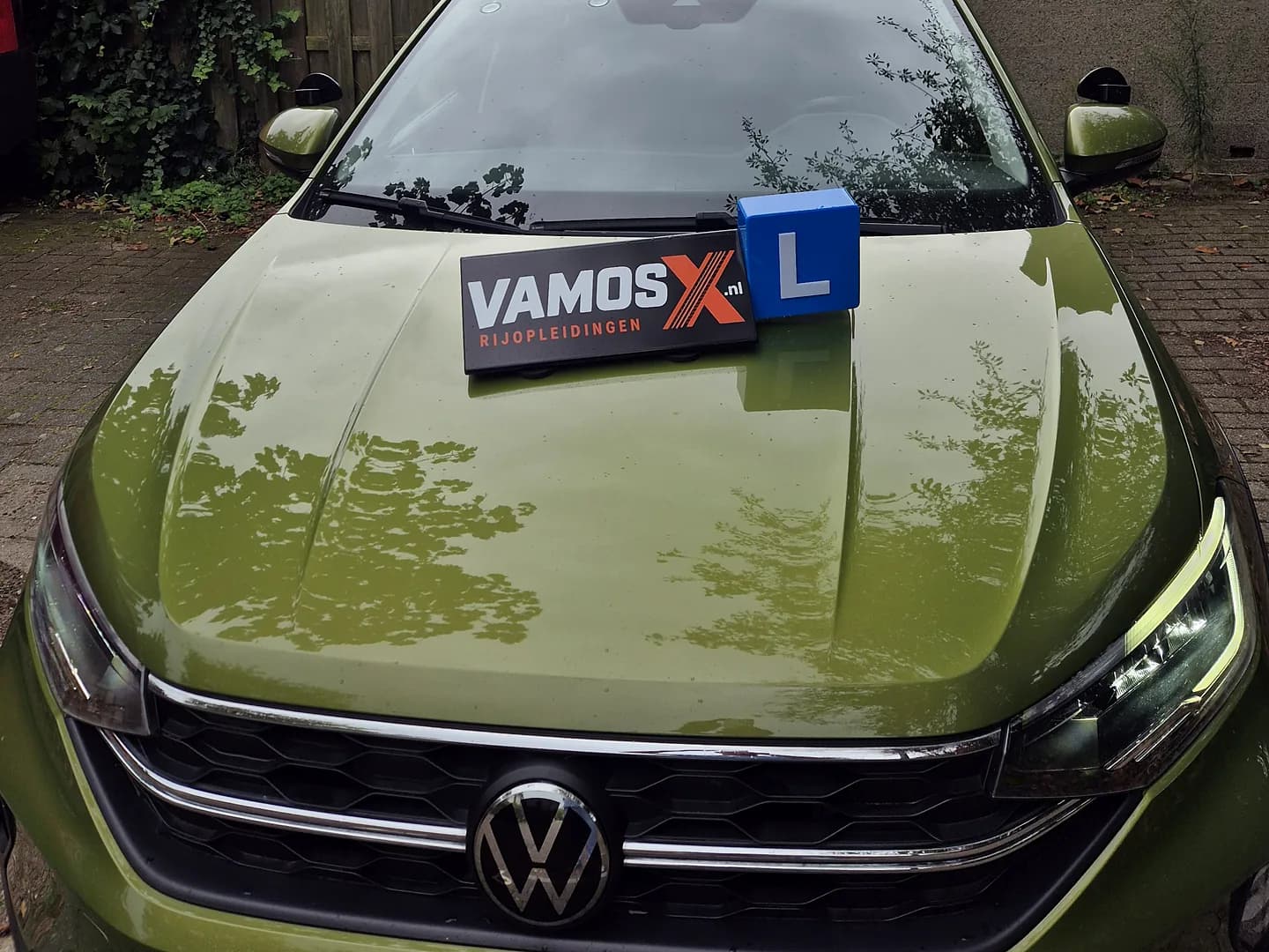 Voorkant van onze groene Volkswagen schakel-lesauto, met het VamosX Rijopleidingen-dakbord en L-blok op de motorkap